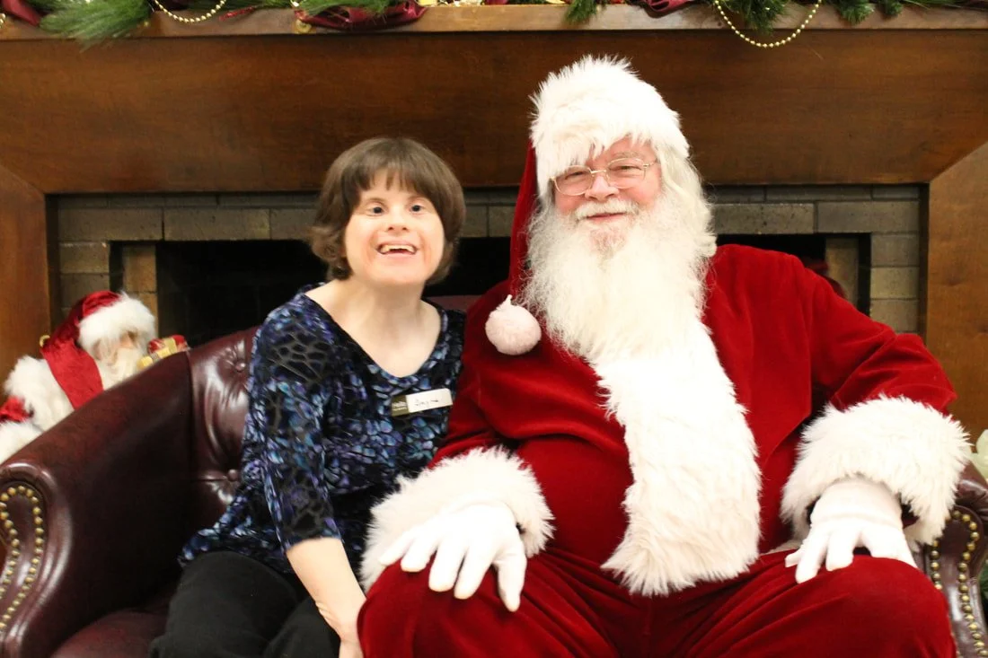 A young girl sitting next to Santa Claus, who is dressed in a red suit with white fur trim, near a fireplace decorated for Christmas.