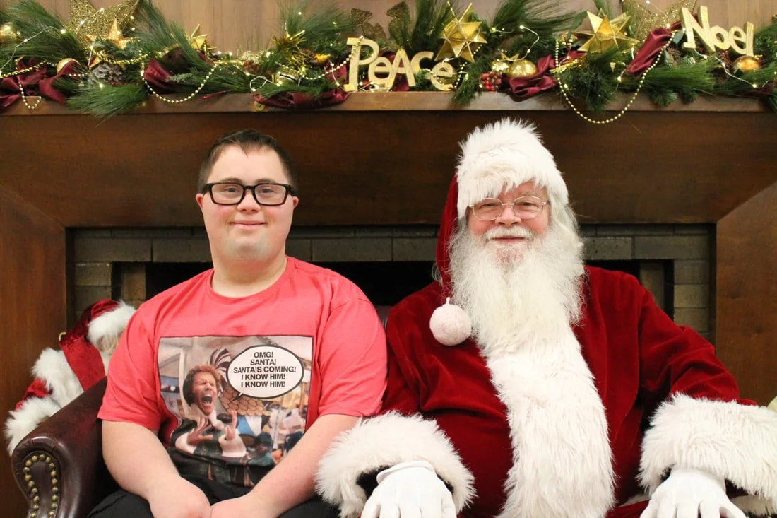 A young man sitting next to Santa Claus in front of a decorated fireplace with Christmas garland, gold stars, and holiday signs reading 'Peace' and 'Noel'.