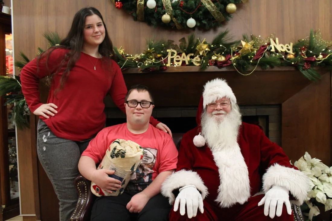 A young woman and a young man sitting with Santa Claus in a Christmas setting with holiday decorations and a wreath in the background.