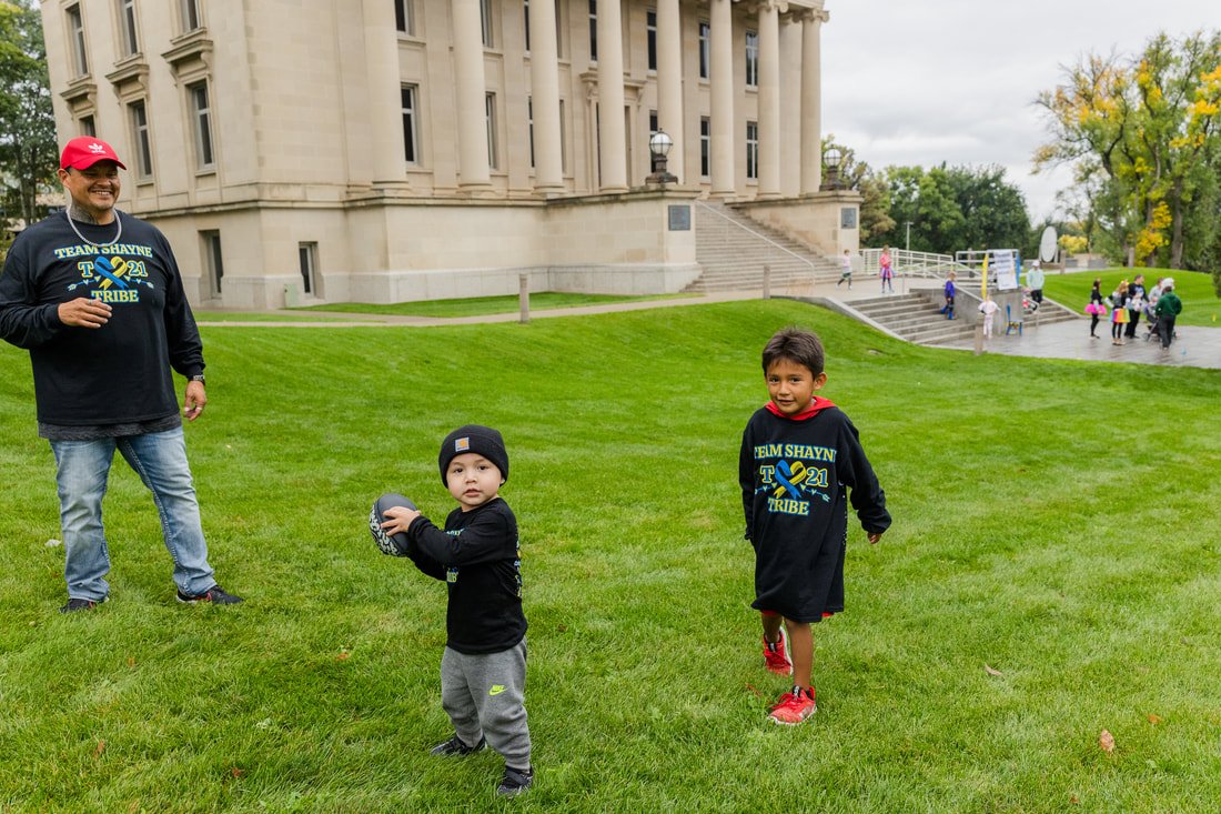 Three children and one adult standing on a green lawn in front of a large, historic building with columns. The children are wearing black shirts with blue and yellow text, and the adult is wearing a similar shirt and a red cap. One child is holding a