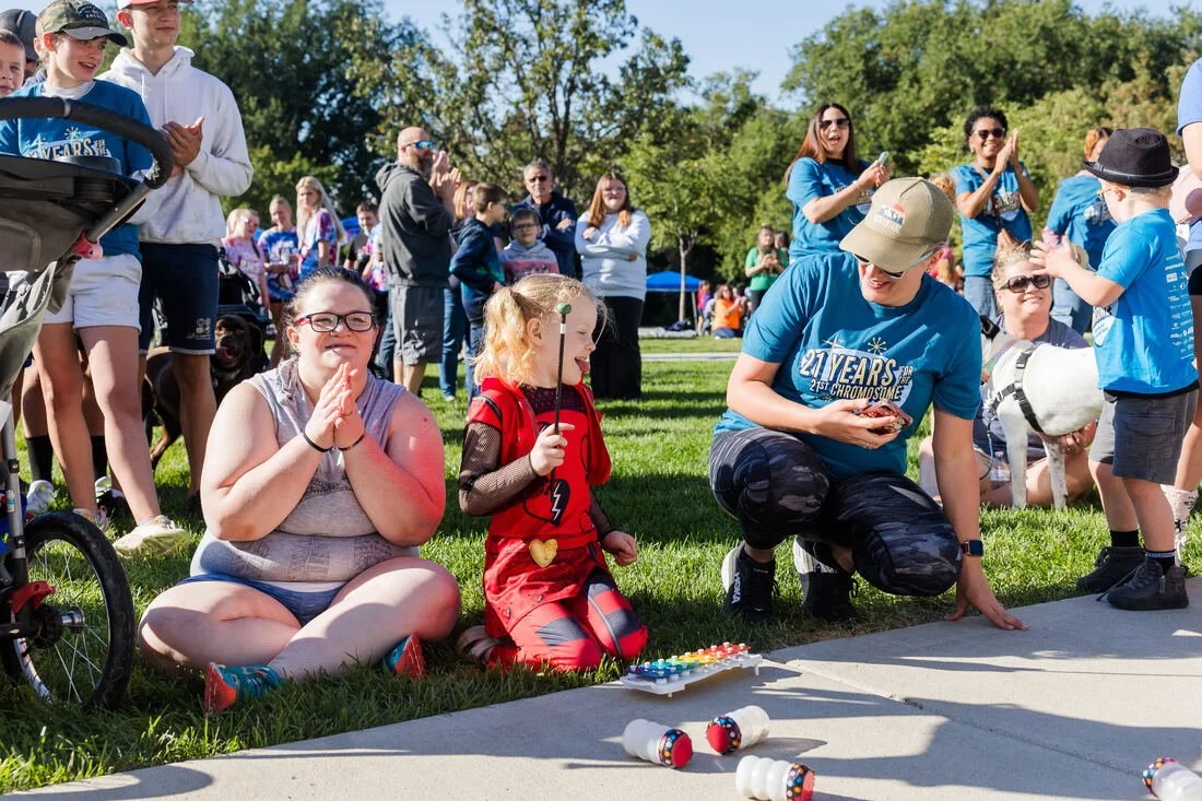 Group of children and adults outdoors at a community event in a park. Two girls are sitting on the grass, one clapping and another holding a small object. A man is kneeling nearby with a smartphone, and a young boy is interacting with small robots on