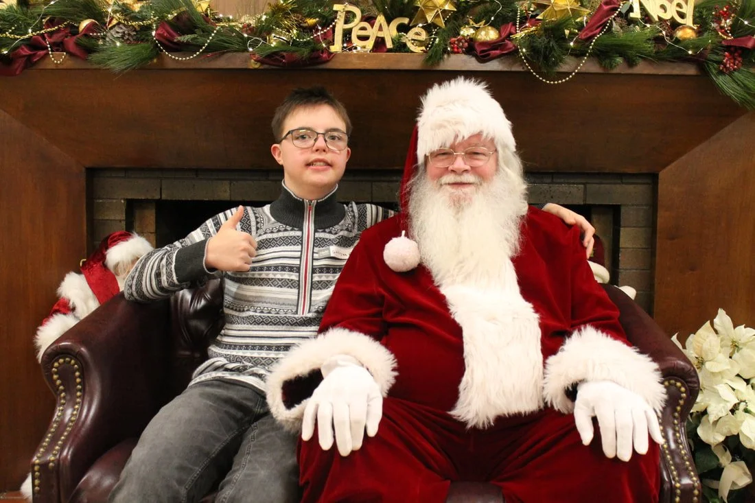A young boy sitting next to Santa Claus in front of a fireplace decorated with Christmas garland and ornaments; the boy is giving a thumbs up and smiling.