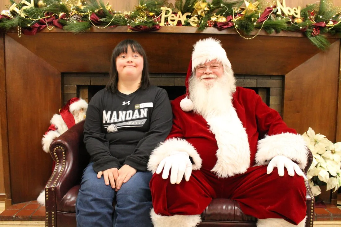 A young person sitting on Santa Claus's lap during a Christmas celebration. Santa is dressed in a red suit with white fur trim, wearing glasses, and has a long white beard. The background has holiday decorations including pine branches, gold ornament