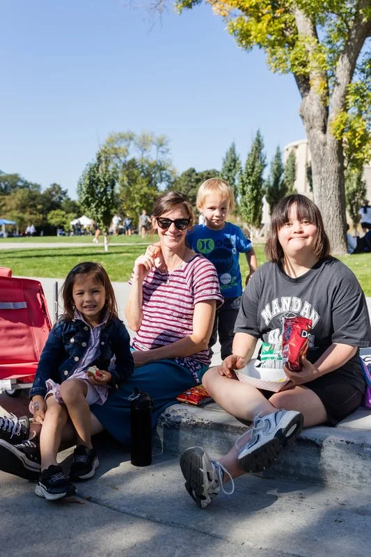 Four children sitting outdoors on a concrete ledge, eating snacks, with trees and a park in the background.