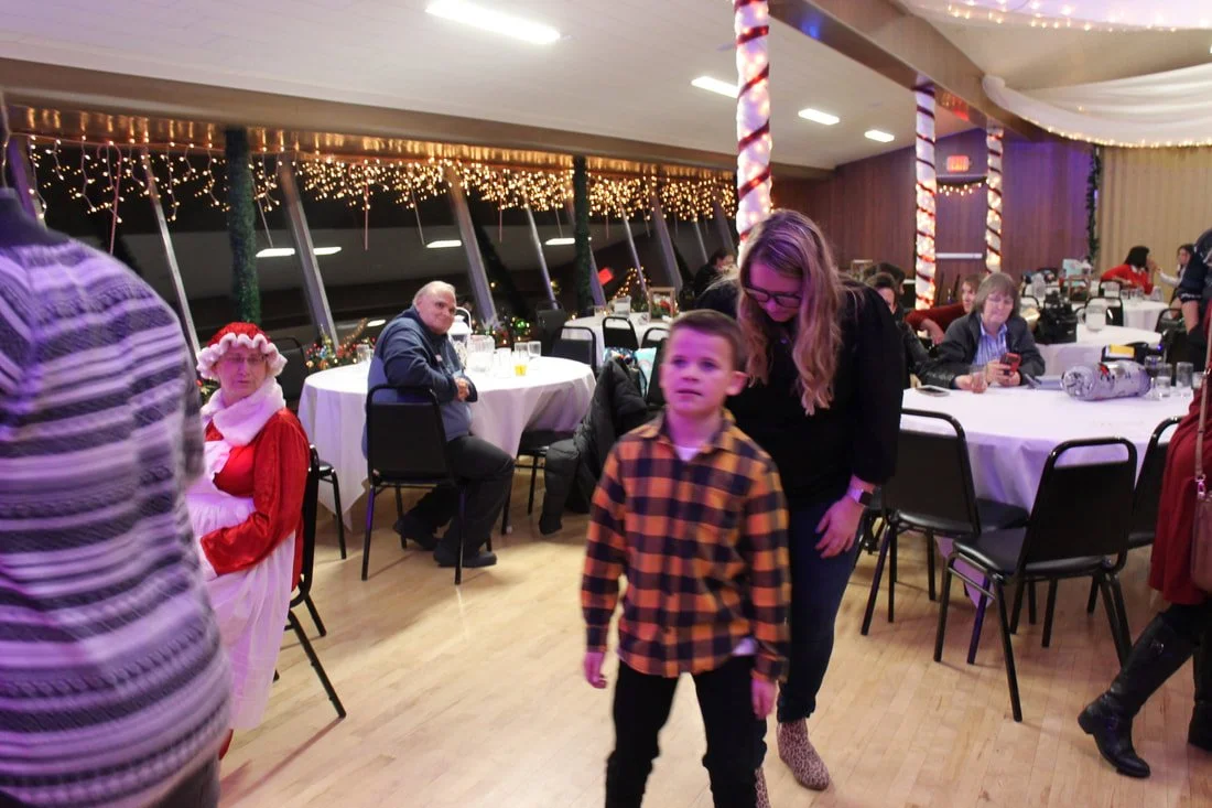 People at a Christmas party with festive decorations, including wrapped candy pole, string lights, and Christmas-themed costumes, seated at round tables in a decorated indoor venue.