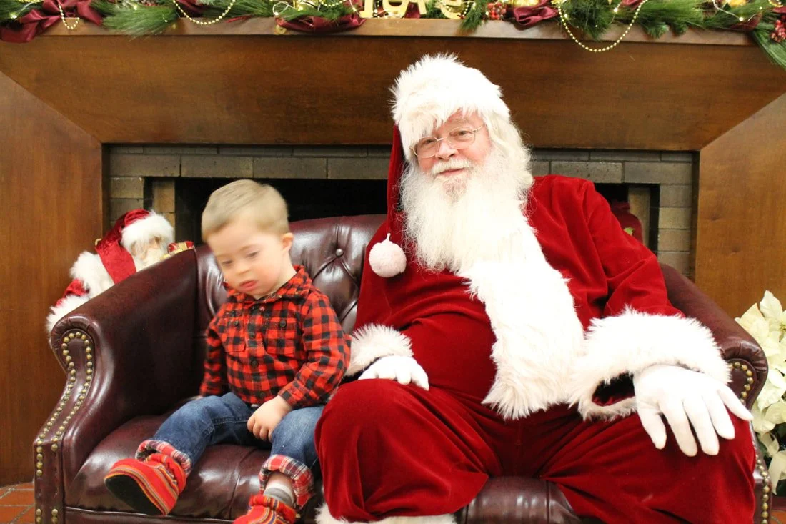 A young boy sitting next to Santa Claus on a leather couch, with a fireplace and Christmas decorations in the background.