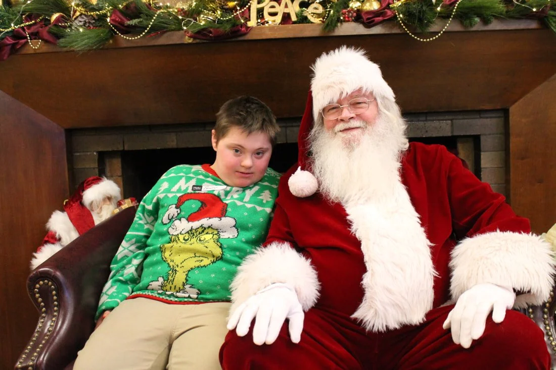 A young boy sitting next to Santa Claus, who is dressed in a red velvet suit with white fur trim, in front of a decorated fireplace with Christmas garland, ornaments, and a 'Peace' sign.