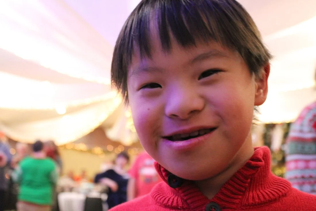 Close-up of smiling young boy with dark hair wearing a red sweater at a festive indoor event, with blurred people and string lights in the background.