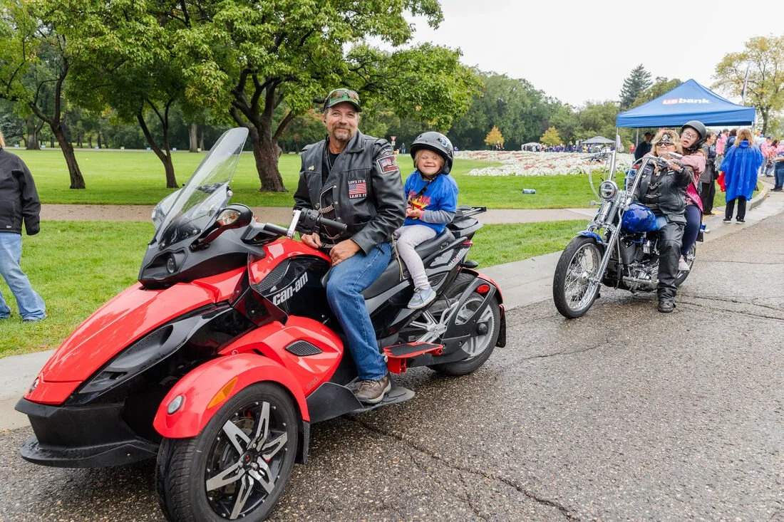 Man and young girl on a red Can-Am three-wheeled motorcycle, woman and young girl on a blue motorcycle, all wearing helmets, in a park with trees and a crowd in the background.
