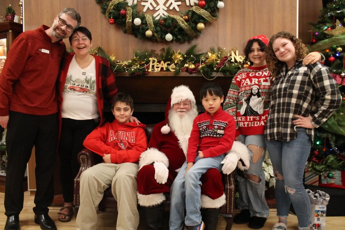 Family and staff gather around Santa Claus for Christmas photo in front of decorated fireplace and Christmas tree.