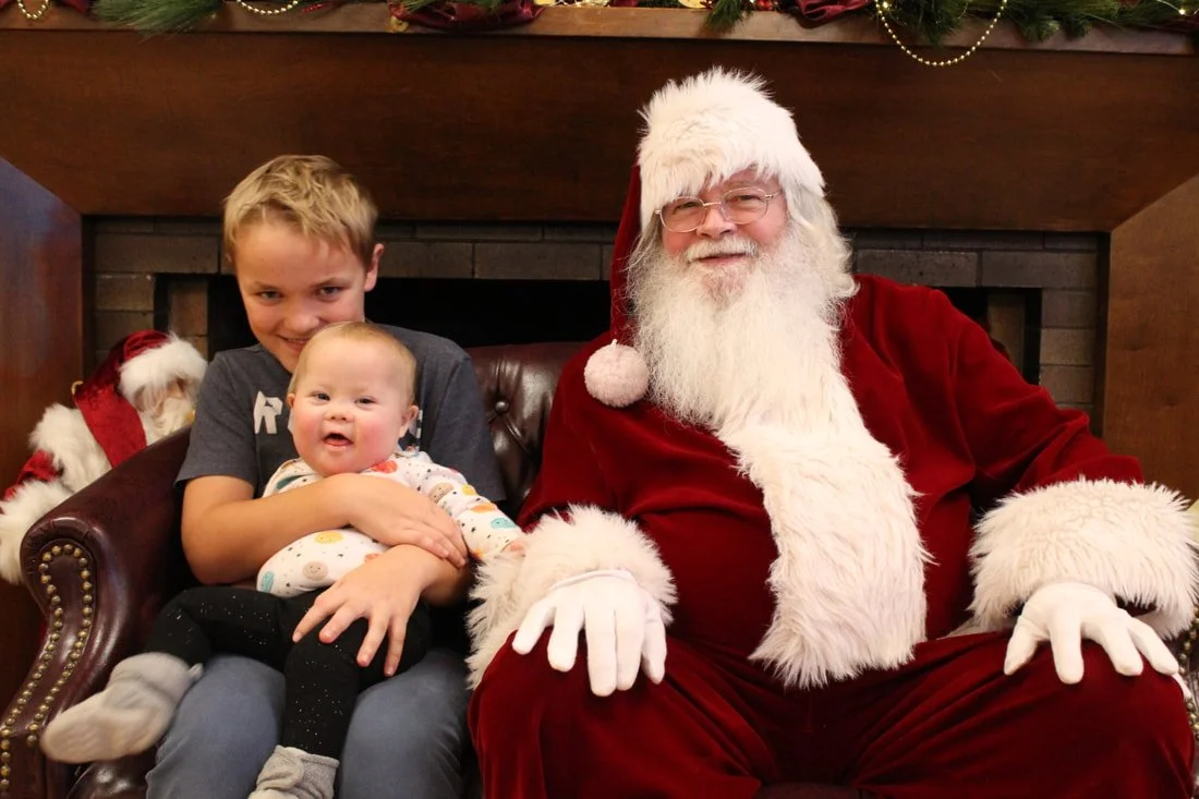 A young boy and a baby sitting next to Santa Claus, who is dressed in a red suit with white fur trim, wearing glasses, and smiling. The children are in front of a fireplace decorated with Christmas garland.