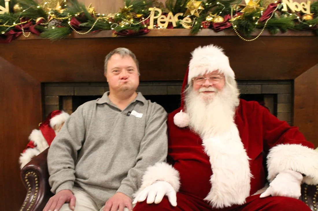 A man making a pouty face sitting next to Santa Claus in a cozy fireplace setting with holiday decorations.