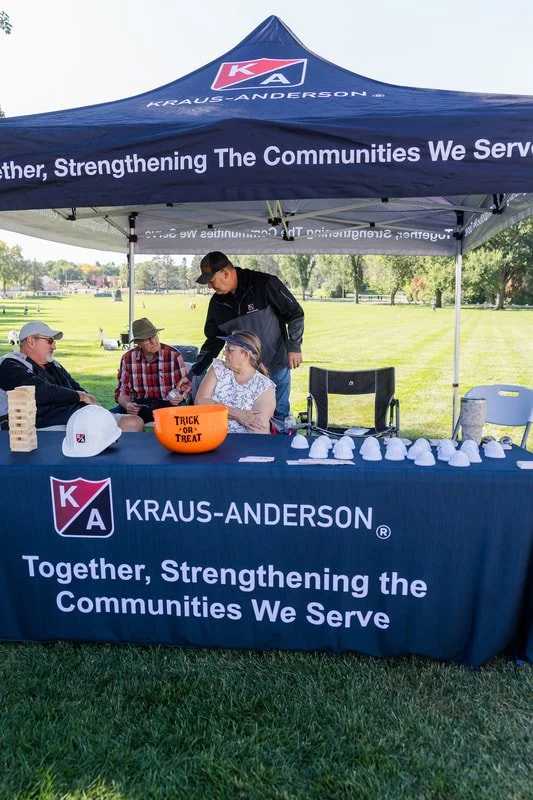 Kraus-Anderson booth at outdoor community event, with a table displaying white lots and a sign for Trick or Treat, under a blue canopy with the slogan "Together, Strengthening the Communities We Serve."