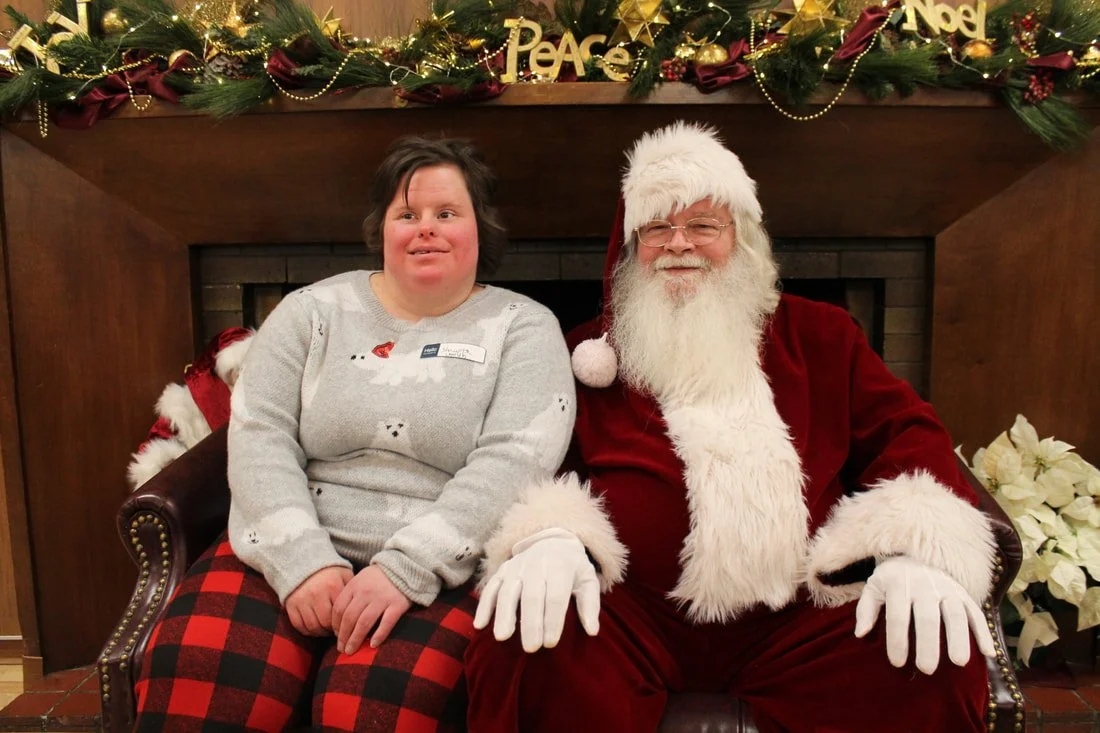 A young woman with a disability and Santa Claus sitting together on a leather chair in front of a Christmas decorated fireplace.