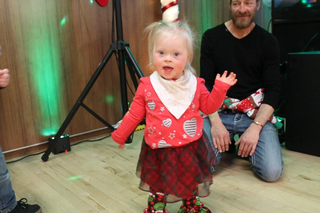 A young girl with blonde hair, wearing a red Christmas sweater with heart and star patterns, a plaid skirt, and festive holiday socks, is dancing and smiling at a Christmas party, with an older man kneeling behind her, smiling.