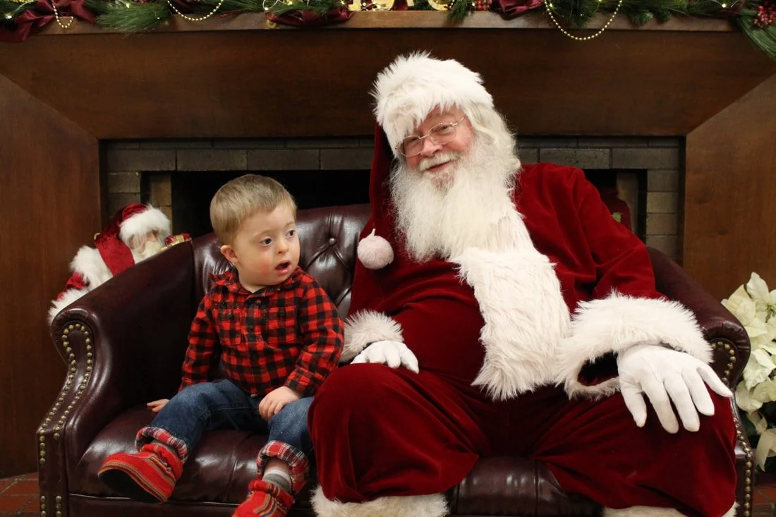 A young boy sitting next to Santa Claus, both seated on a leather chair, with Christmas decorations above them and a fireplace in the background.