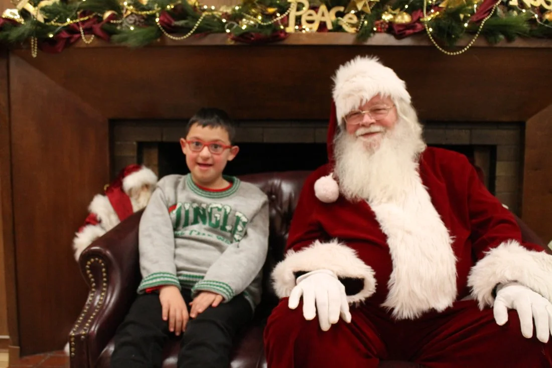 A young boy with glasses sitting next to Santa Claus on a leather chair in front of a decorated fireplace, with a stocking hanging behind them.