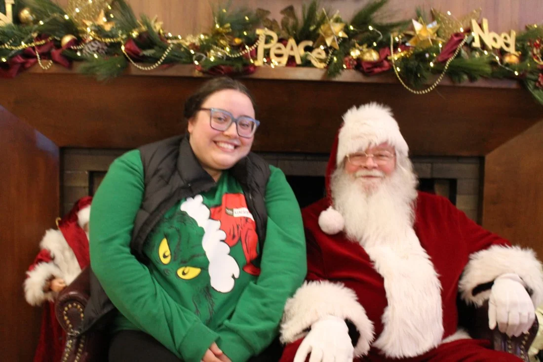 A person wearing a green festive shirt with a Grinch design and glasses sitting next to Santa Claus in a red suit with white fur trim, both smiling, with holiday decorations and garland over a fireplace in the background.