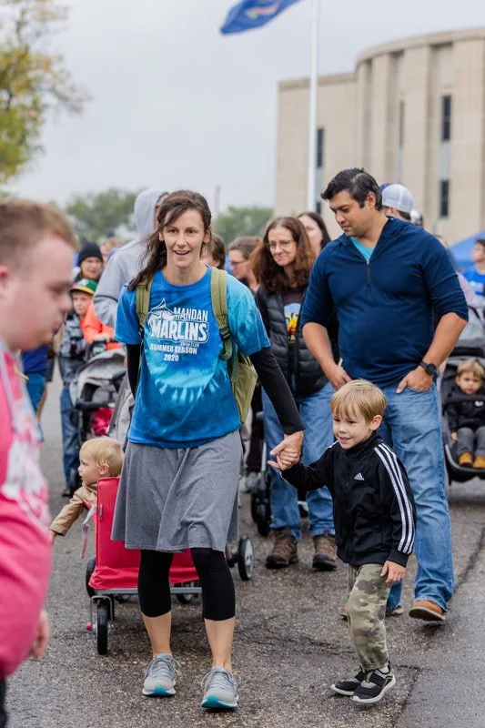 A woman in a blue Marlins summer season 2020 shirt holding hands with a young boy in a black jacket, walking outdoors among a group of people, with some children in strollers and a building in the background.