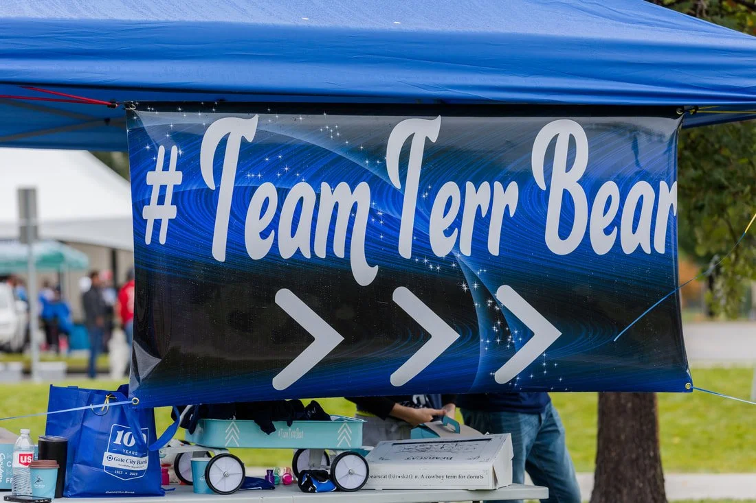 A blue banner with the text '#TeamTerr Bear' and arrows, hanging under a blue canopy at an outdoor event.