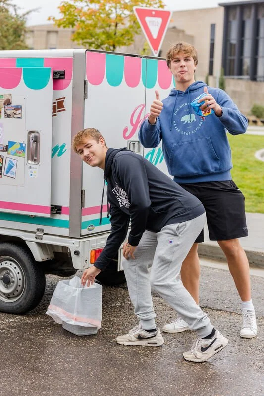 Two teenage boys in casual clothing by an ice cream vending cart. One is bending down picking up a plastic bag, and the other is standing with a thumbs-up sign holding a drink.