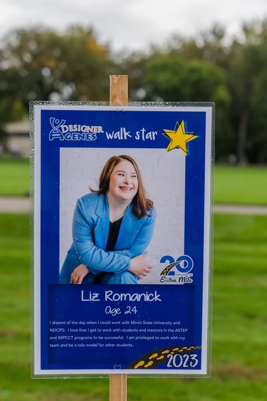 Sign honoring Liz Romanick, age 24, as a 'walk star' at the 2023 Extra Mile event, featuring her photo, a large yellow star, and celebratory graphics against a park background.