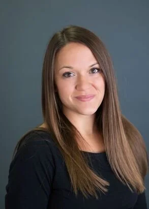 A woman with long brown hair wearing a black top, smiling slightly, standing against a blue background.