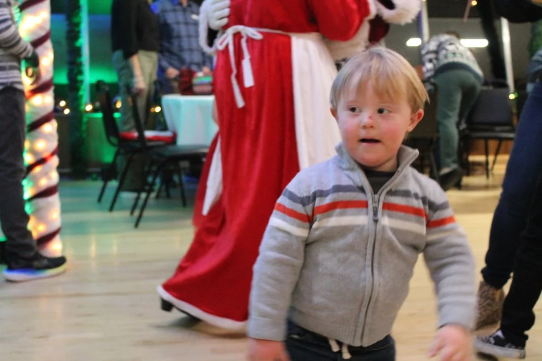 A young boy with blond hair and a striped gray and red sweatshirt standing indoors at a holiday event.