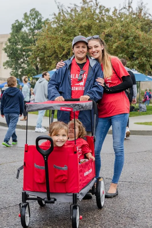 Two young women standing behind a red stroller with two laughing children inside at an outdoor event, with other people in the background.