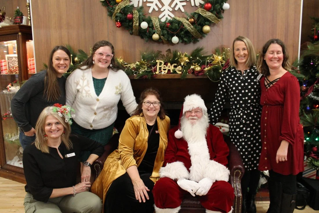 Group of seven women and Santa Claus sitting and standing in front of a decorated Christmas mantle with a large holiday wreath and garland. The women are smiling, dressed in festive clothing, and are positioned around Santa who is seated in the cente
