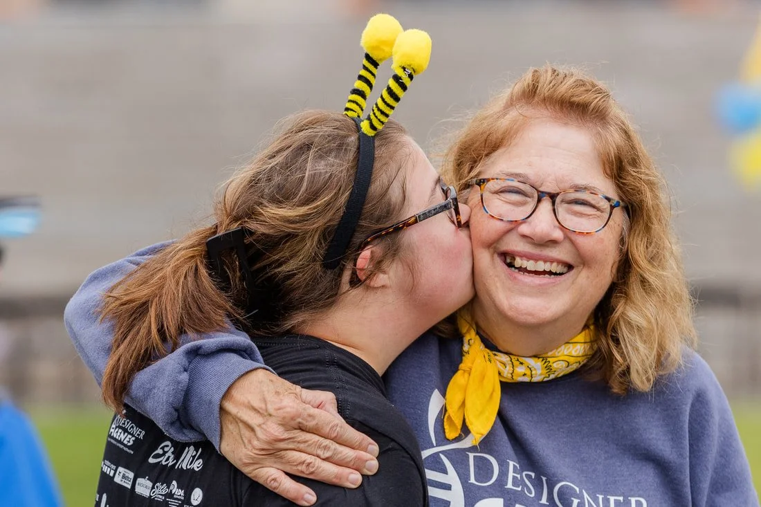 A young woman dressed in a bee costume kisses an older woman with glasses and a yellow bandana around her neck, both smiling.