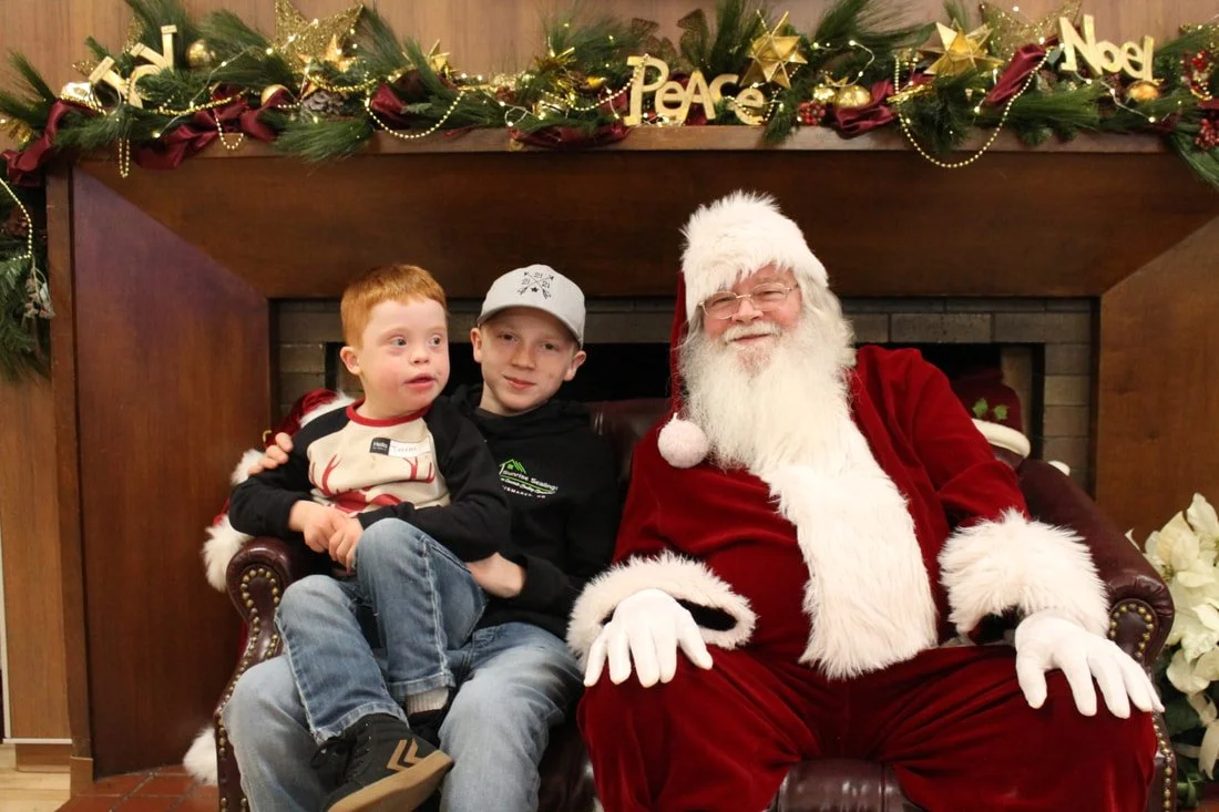 Two boys sitting with Santa Claus in front of a decorated fireplace with Christmas ornaments and gold 'Peace' and 'Noel' signs.