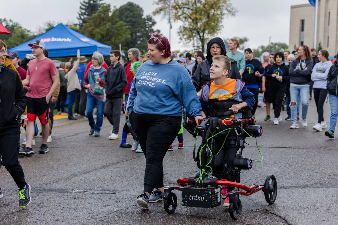 A diverse group of people participating in an outdoor event, with a woman in a blue sweatshirt holding a young boy on a walker equipped with various devices, surrounded by others under tents and trees.