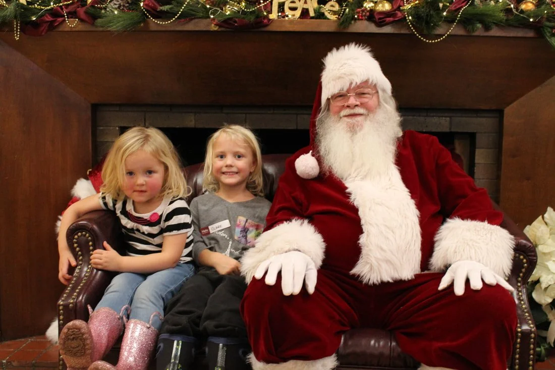 Two young girls sitting with Santa Claus in front of a Christmas fireplace decorated with ornaments and lights.