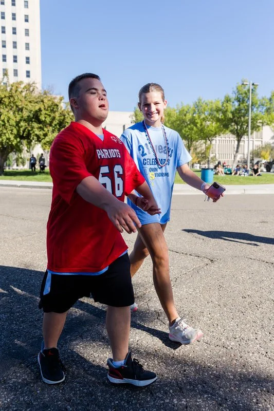Two kids, a boy in a red sports jersey and a girl in a blue T-shirt, walking outdoors on a sunny day with trees and buildings in the background.