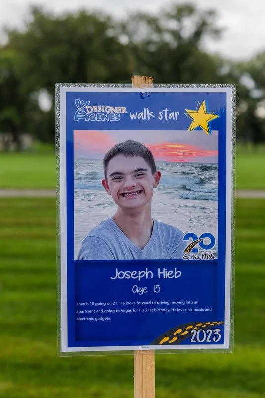 A sign resembling a school or campaign sign with a photo of a boy smiling at the beach at sunset. The sign reads "Designer Genes Walk Star," 