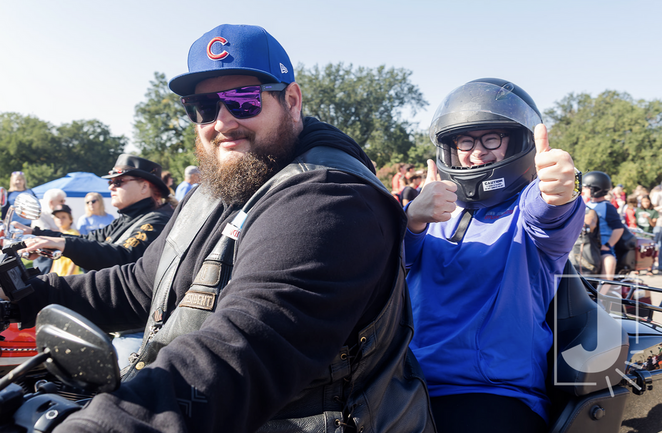A man with a beard and sunglasses riding a motorcycle, and a young girl behind him wearing a helmet and giving a thumbs-up at an outdoor event with other people and trees in the background.