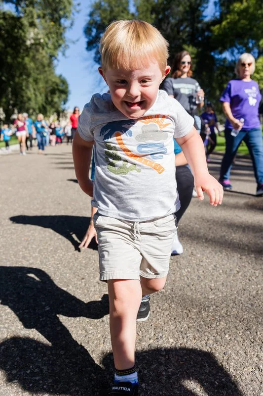 Young boy running outdoors during a group walk or race, with a big smile, dressed in a light-colored t-shirt and shorts, on a sunny day with trees and people in the background.