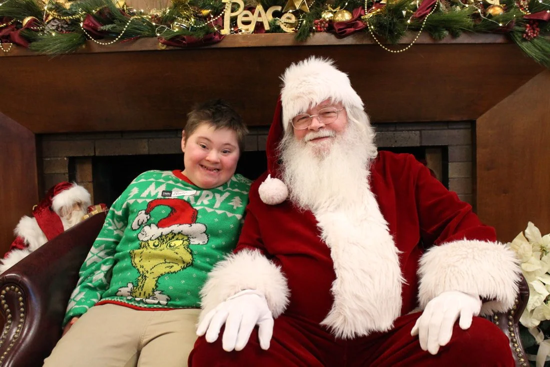 A young boy sitting next to Santa Claus in front of a festive fireplace decorated with Christmas ornaments, greenery, and a 'Peace' sign, at a holiday event.