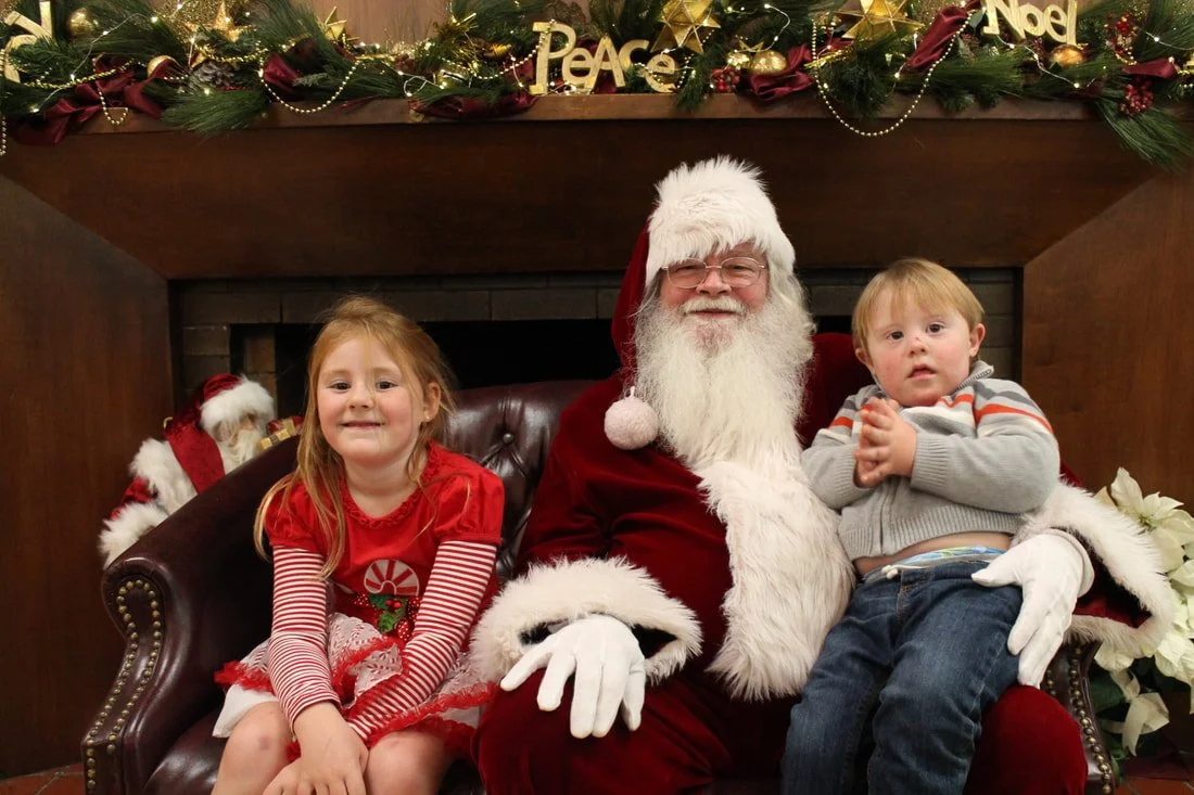 A man dressed as Santa Claus sitting on a leather chair with two children, a girl and a boy, in front of a Christmas decorated fireplace with garland, ornaments, and holiday signs.