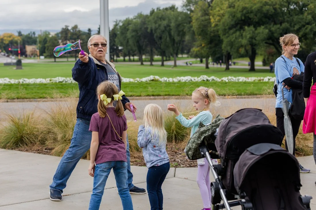 An older woman blowing bubbles for three young girls in a park, with two women standing nearby.