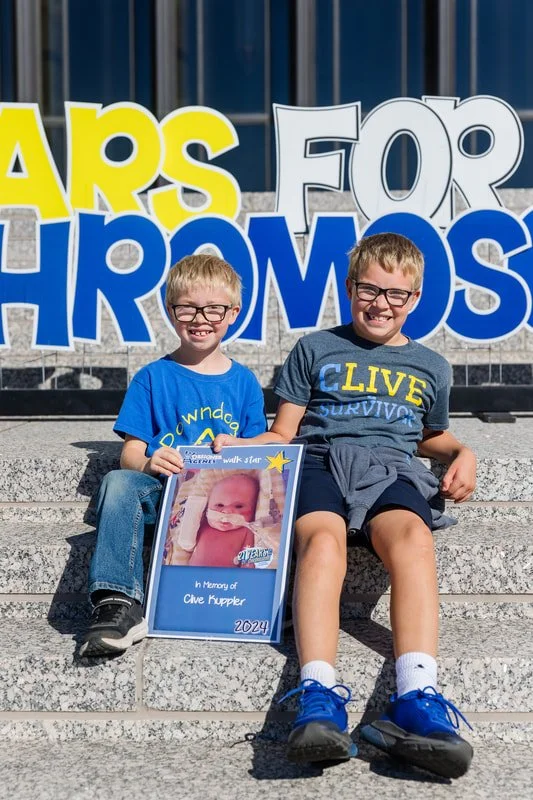 Two young boys sitting on steps outside, smiling, with a colorful sign in the background that reads 'Cards for Chromosomes.' One boy holds a framed memorial picture of a baby with a note saying 'In Memory of Clive Kuppler, 2024'.
