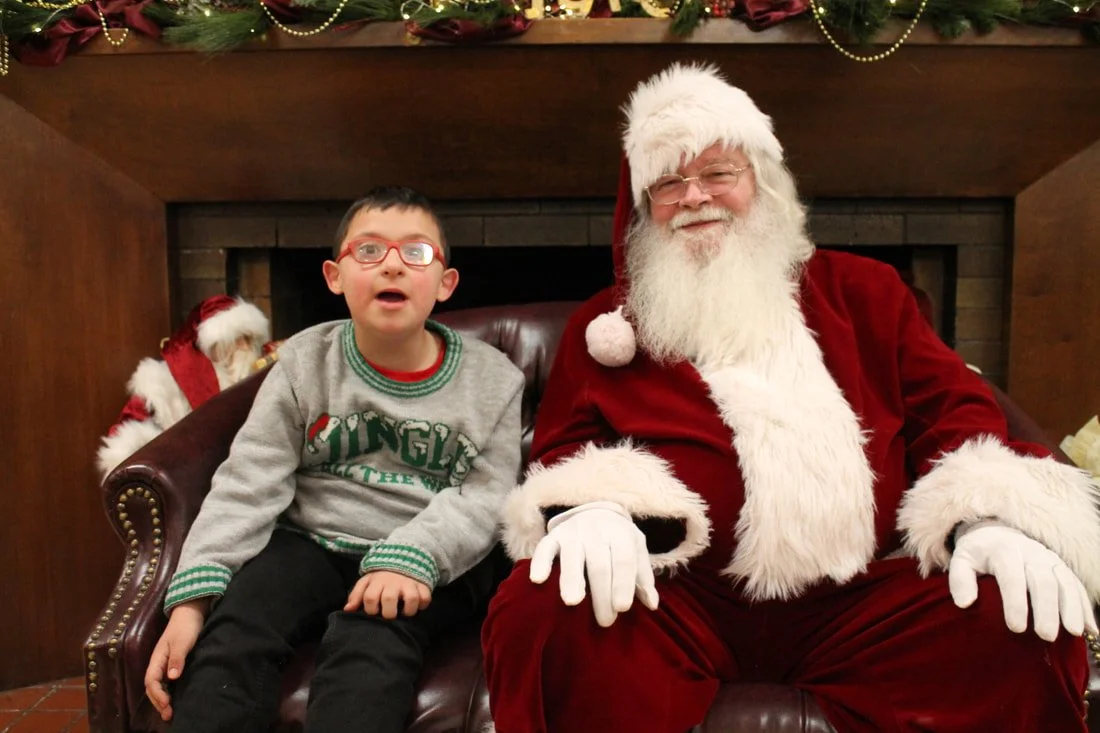 A boy with glasses sitting next to Santa Claus, who is dressed in a red suit with white fur trim, in front of a decorated fireplace with a Santa figurine behind them.