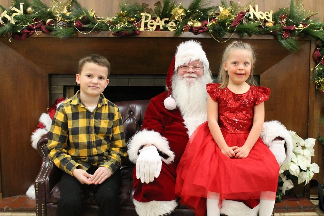 A young boy in a yellow and black checkered shirt and a young girl in a red dress sit with Santa Claus on a decorated Christmas mantle. The mantle is adorned with greenery, gold stars, ribbons, and ornaments. The scene is festive with a Christmas the