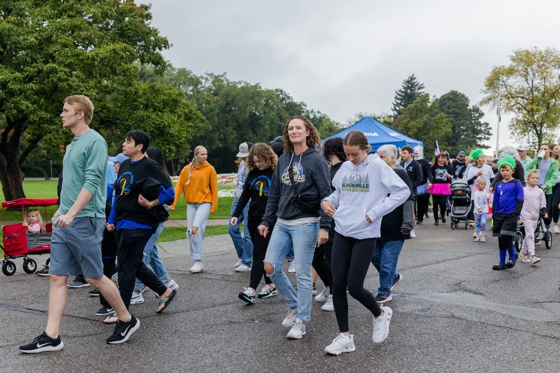 Group of people walking in a park during an outdoor event, some wearing hoodies and jackets, children in strollers, green trees, overcast sky.
