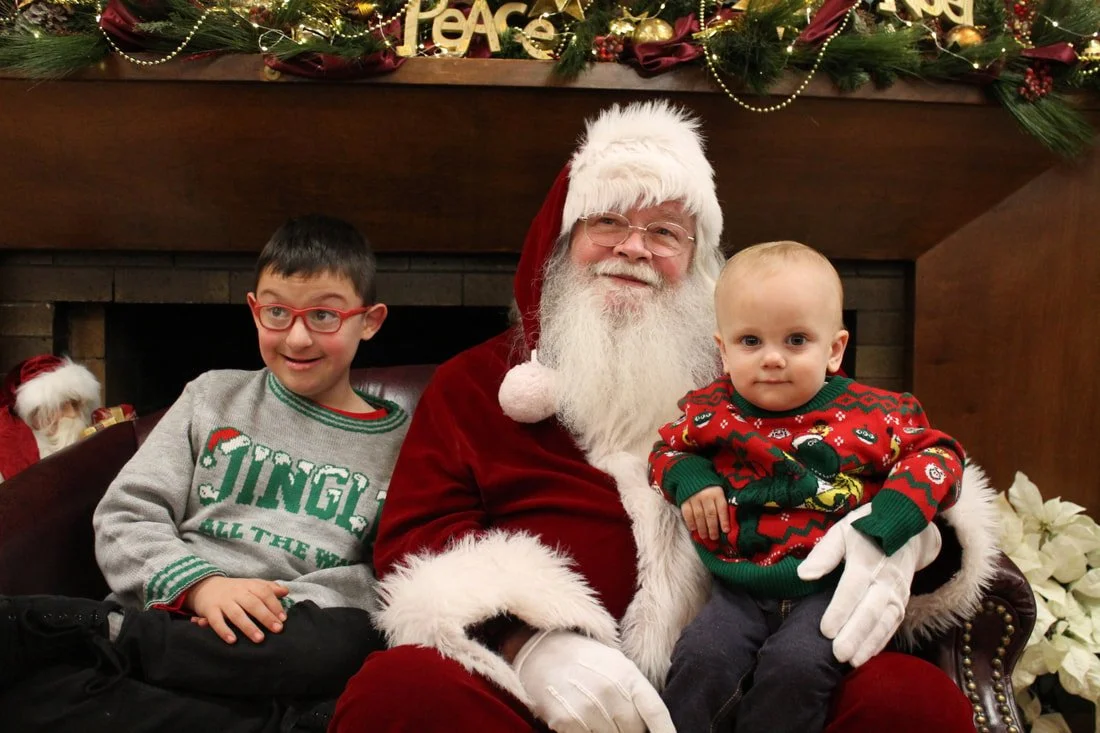 A man dressed as Santa Claus sitting on a couch with two children, one boy with glasses and a Christmas sweater, and a younger child in a holiday sweater, in a festive setting with Christmas decorations and garland.