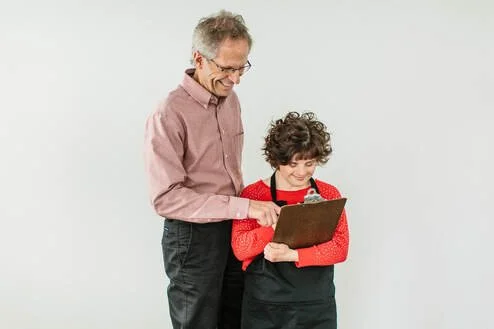 An older man and a young girl looking at a clipboard together, smiling against a plain white background.