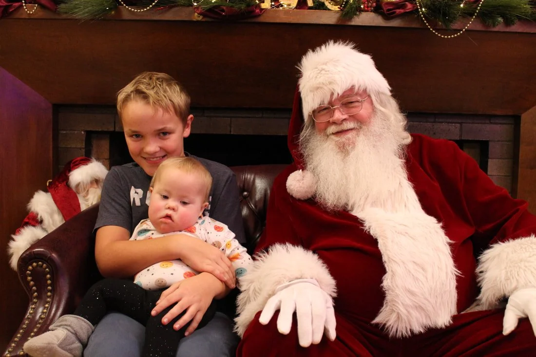 A young boy and a baby sitting with Santa Claus in front of a decorated fireplace with greenery and ornaments, during Christmas.