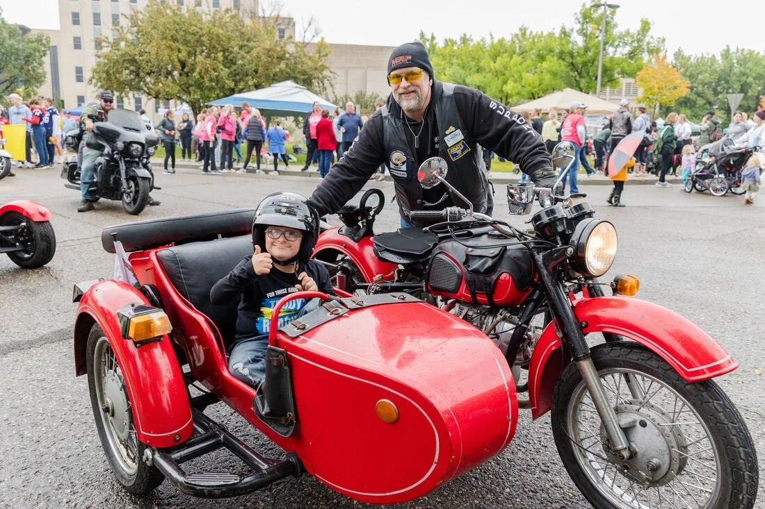 A young boy with glasses and a helmet sitting in a red motorcycle with sidecar, giving a thumbs-up, while an older man in sunglasses and a black jacket stands behind the motorcycle at a busy outdoor event with many people and tents in the background.
