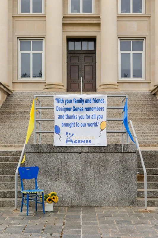 A banner on stairs reads, "With your family and friends, Designer Genes remembers and thanks you for all you brought to our world!" The stairs have a blue chair and a flower pot with sunflowers, with yellow and blue ribbons on the sides.